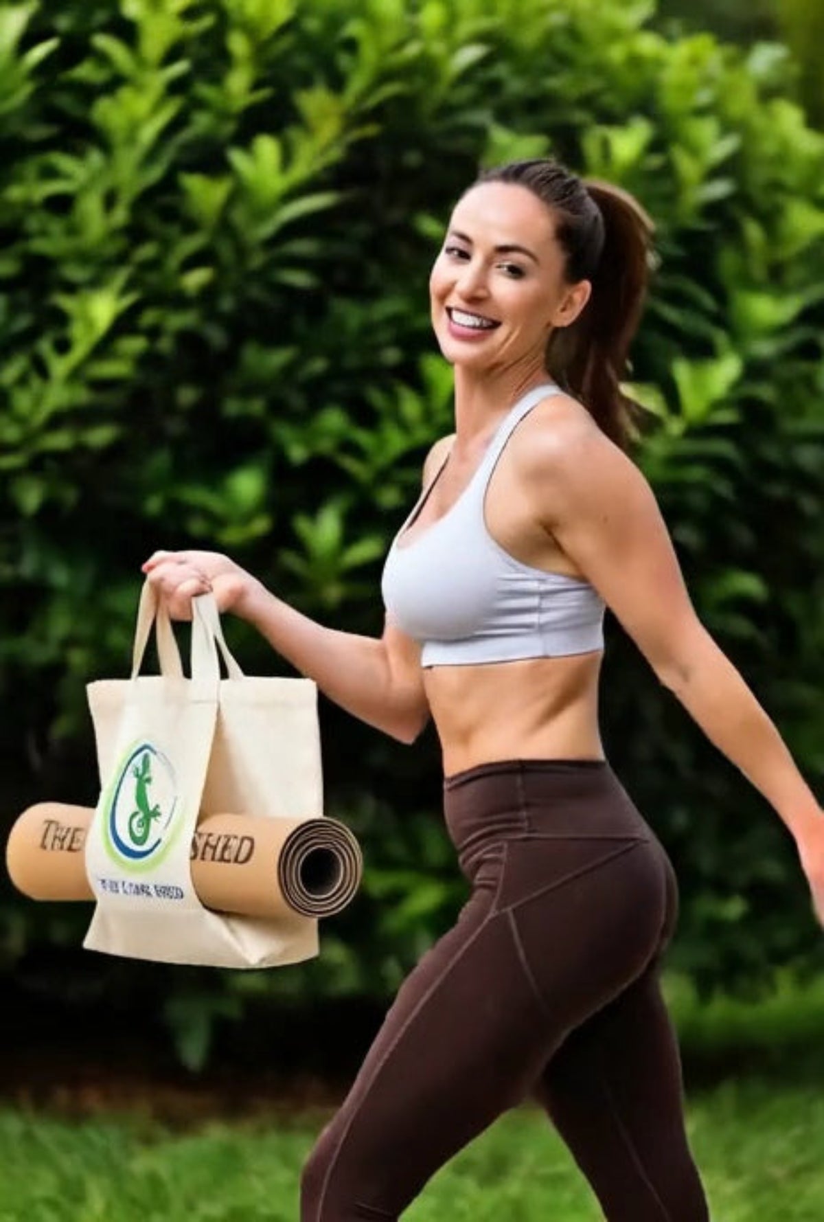 Woman in athletic wear holding a yoga mat and a bag with greenery in the background